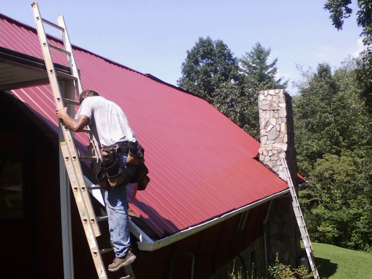 Roofer on a ladder beside a red metal roof and stone chimney — Soffit & Fascia Repair by Roofing Brothers in Lemont Furnace